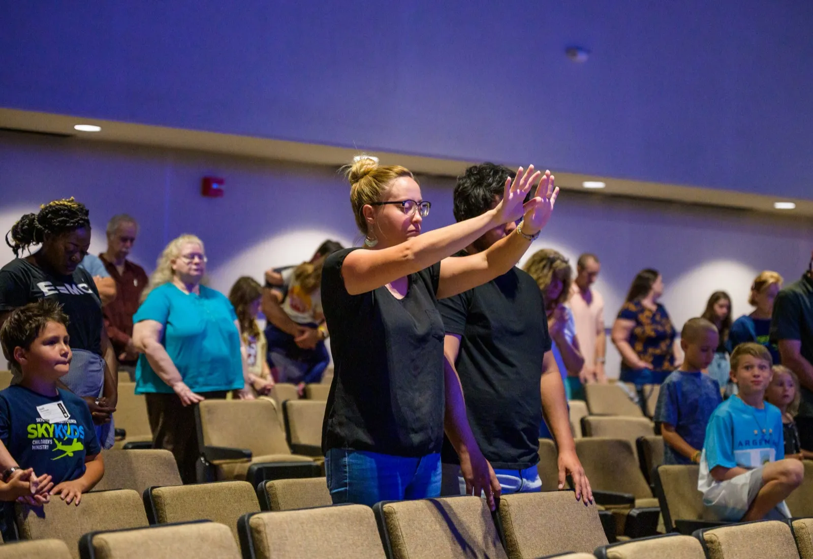 Skycrest worshippers standing and raising their hands during a service, with children and adults together in the sanctuary