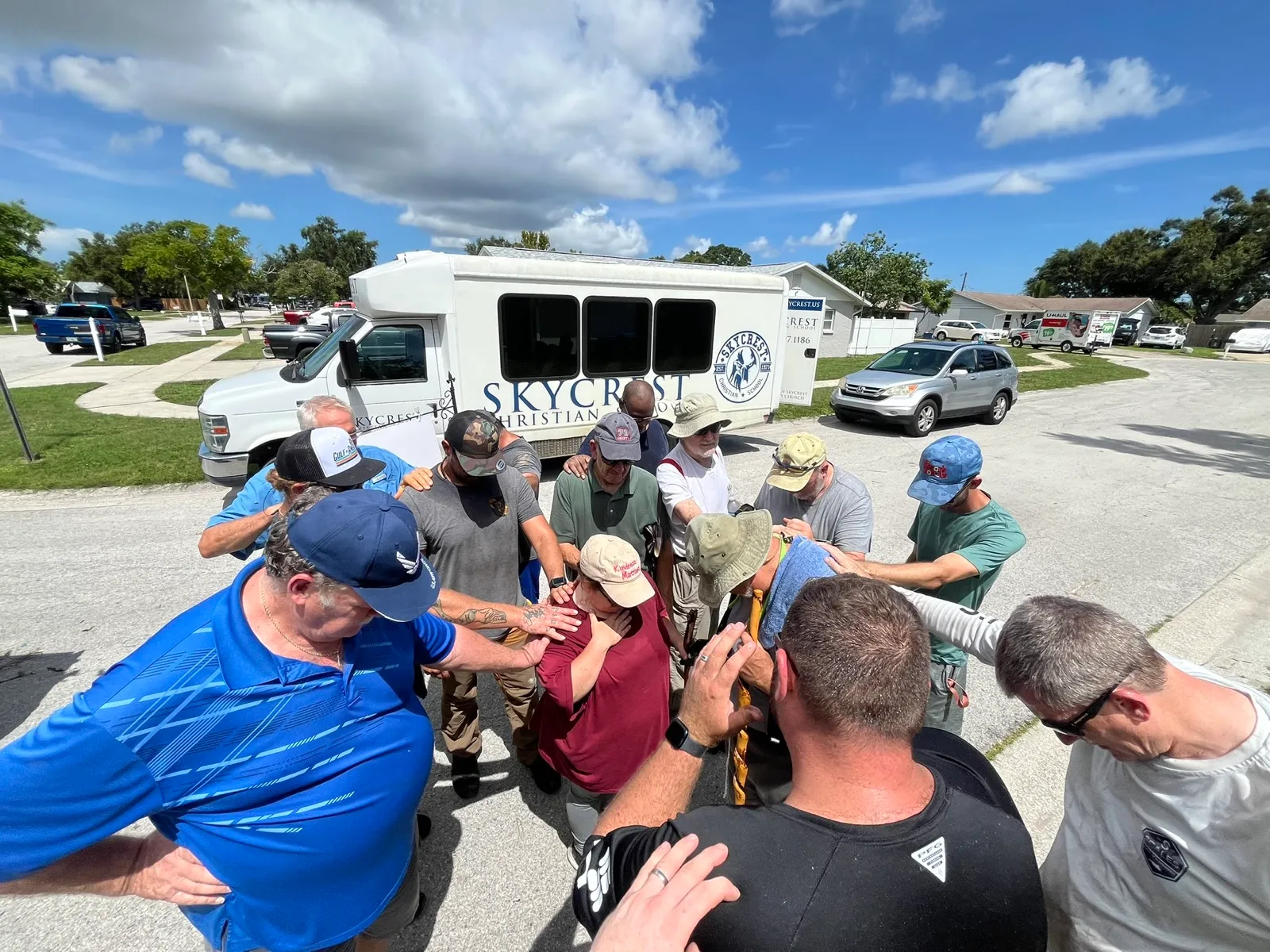 A group gathered in a circle in the church parking lot with hands on one another's shoulders and heads bowed in prayer