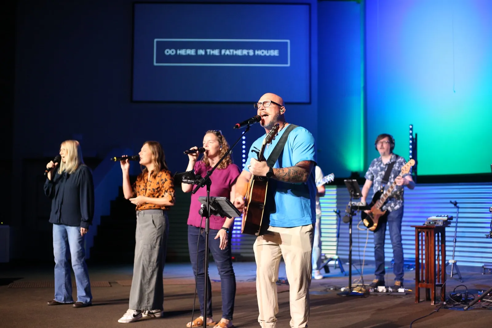 The Skycrest worship team mid-song — the worship leader singing at the mic with an acoustic guitar, three vocalists beside him and a bassist stage right